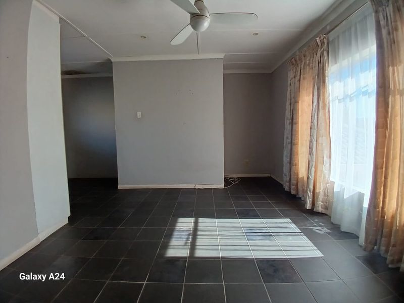 An empty living room featuring dark tiled flooring, a ceiling fan, and large windows with curtains, offering a blank canvas for interior design.