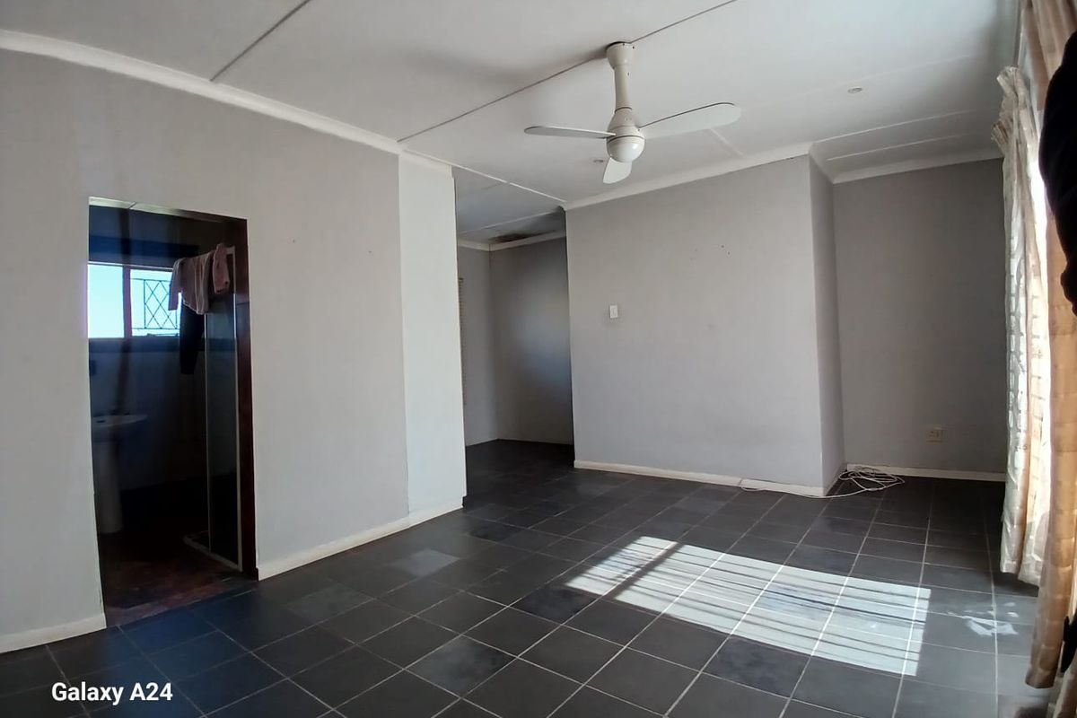A spacious, empty living area featuring dark tiled flooring, a ceiling fan, and neutral grey walls, with an adjoining room visible.