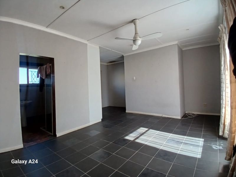 A spacious, empty living area featuring dark tiled flooring, a ceiling fan, and neutral grey walls, with an adjoining room visible.