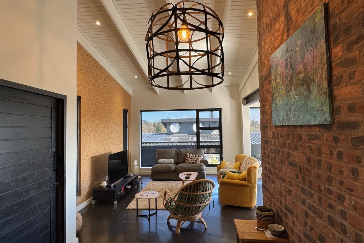 Spacious living room featuring a vaulted ceiling, exposed brick wall, cork feature wall, and polished concrete flooring with large windows providing natural light.