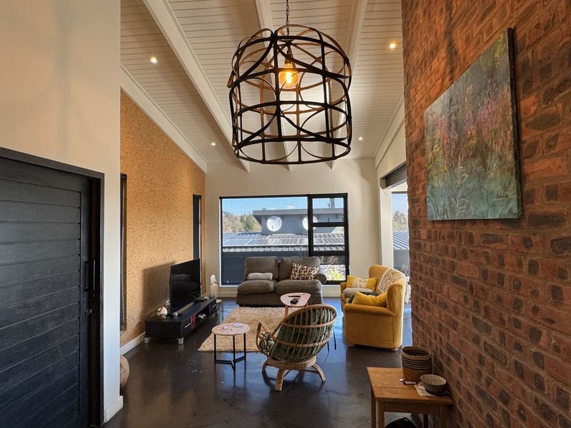 Spacious living room featuring a vaulted ceiling, exposed brick wall, cork feature wall, and polished concrete flooring with large windows providing natural light.