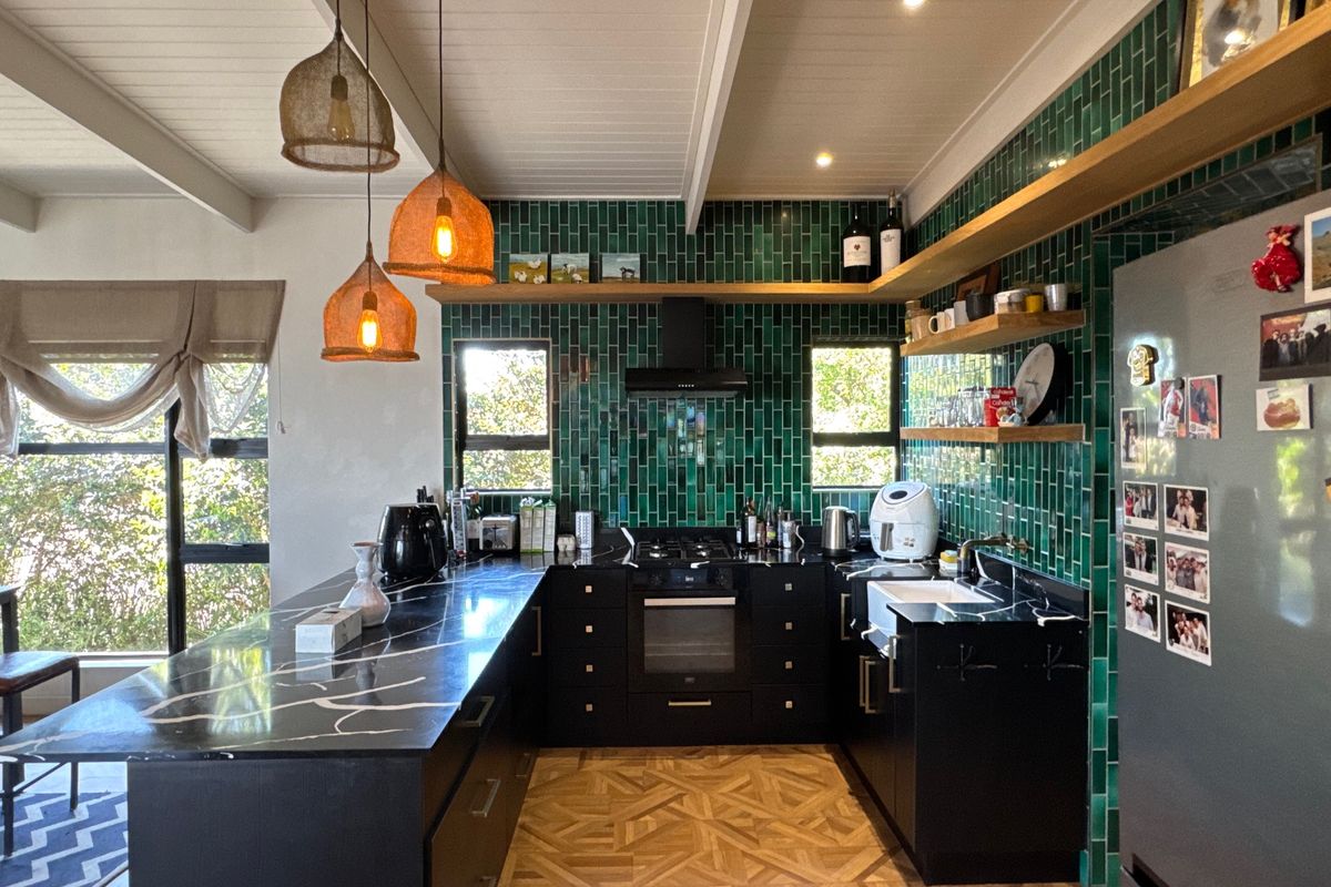 Modern kitchen featuring striking green subway tile backsplash, black marble-effect countertops, farmhouse sink, built-in oven, and warm wooden floating shelves under exposed beam ceilings.