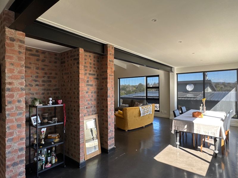 Spacious open-plan living and dining area featuring industrial-style exposed brick columns, steel beams, polished concrete floors, and large windows with natural light.