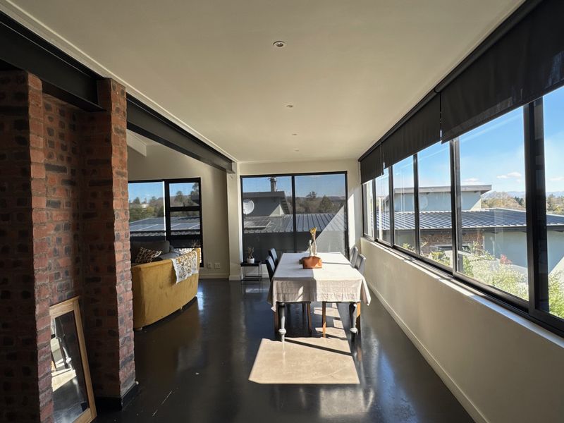 Bright open-plan living and dining area featuring polished concrete floors, exposed brickwork, steel structural beams, and expansive windows with integrated roller blinds.