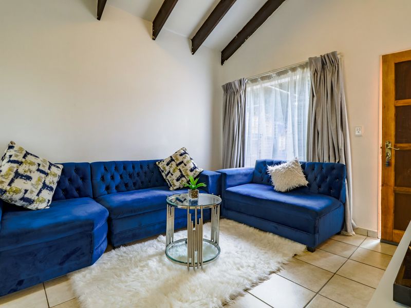 A cozy living room featuring a vaulted ceiling with exposed timber beams, tiled flooring, and a wooden door leading to the exterior.