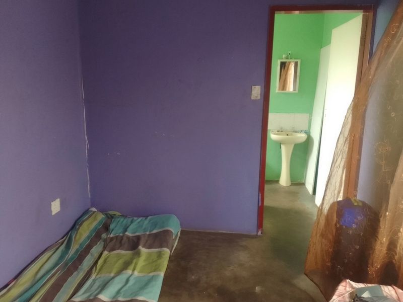 A bedroom with purple walls and concrete flooring, featuring an adjoining bathroom area with a pedestal basin and wall mirror.