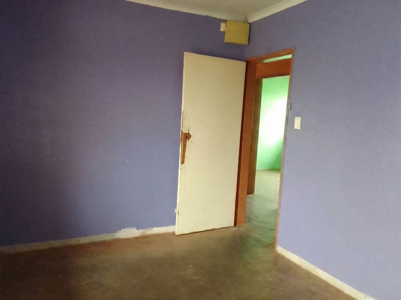An empty bedroom featuring purple walls, a damaged internal door, and exposed concrete flooring, requiring cosmetic updates and repairs.