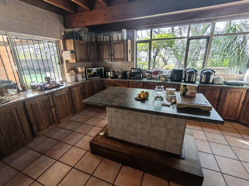 Spacious kitchen featuring warm wooden cabinetry, a central island, tiled flooring, and exposed timber ceiling beams with large windows overlooking the garden.