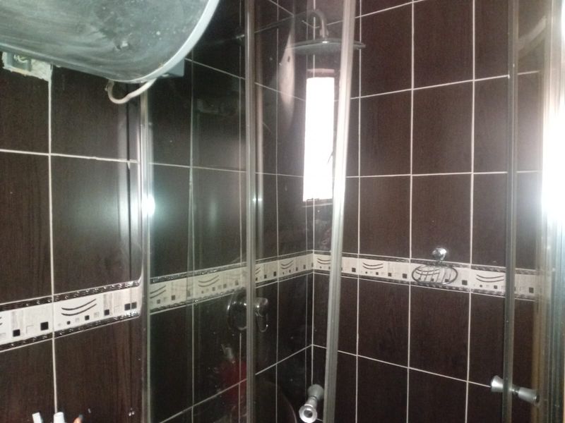 A compact bathroom featuring dark wall tiles, a glass shower enclosure, and an overhead shower fixture with an exposed electric water heater unit.