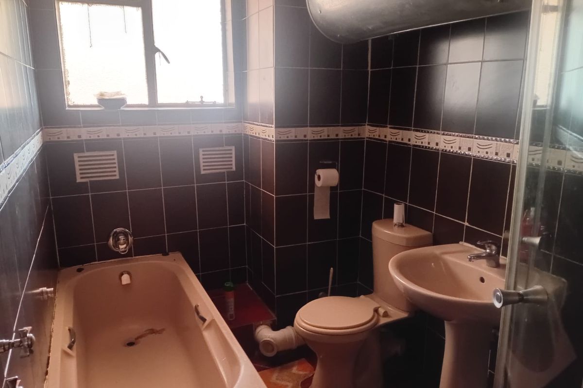 A compact bathroom featuring dark wall tiles, a pedestal basin, toilet, and a bathtub with exposed plumbing and dated fixtures.