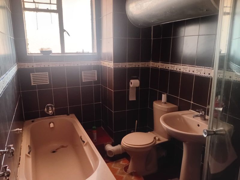 A compact bathroom featuring dark wall tiles, a pedestal basin, toilet, and a bathtub with exposed plumbing and dated fixtures.