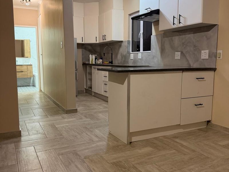 Modern kitchen featuring white cabinetry, stone countertops, and herringbone-patterned floor tiles, with an open-plan layout leading to a hallway and bathroom.