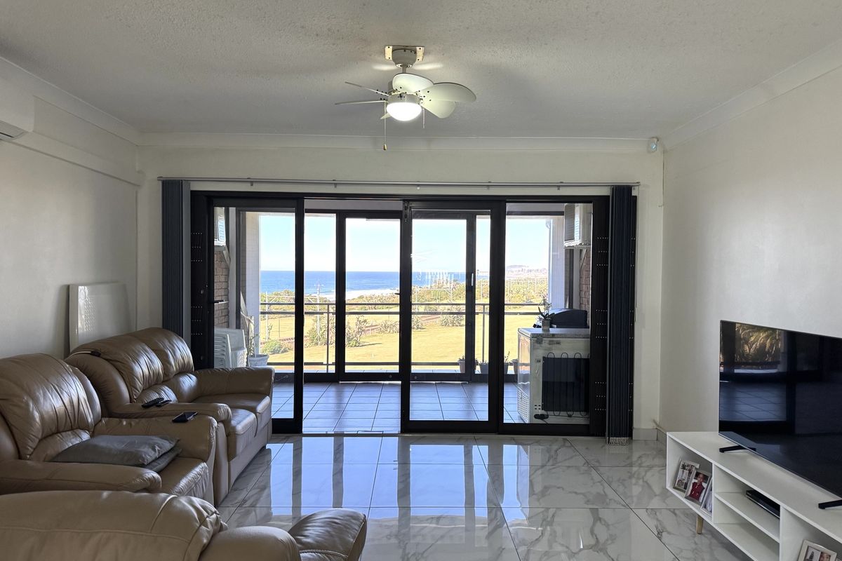 Spacious living room featuring polished marble-look floor tiles, a ceiling fan, and large sliding glass doors opening onto a balcony with scenic coastal views.