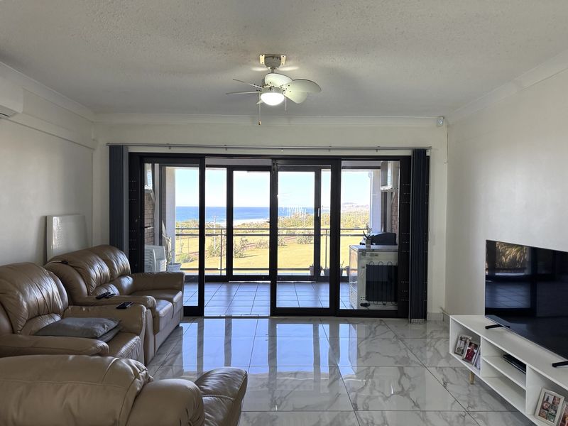 Spacious living room featuring polished marble-look floor tiles, a ceiling fan, and large sliding glass doors opening onto a balcony with scenic coastal views.