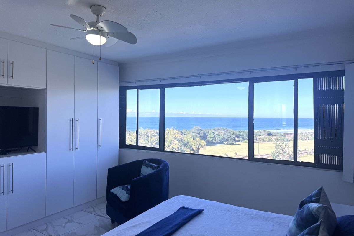A bright bedroom featuring built-in cupboards, a ceiling fan, and large windows offering expansive ocean and coastal views.