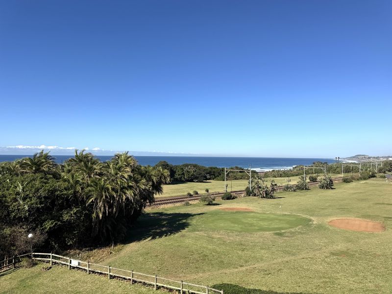 A scenic elevated view overlooking a golf course, railway line, and the coastline with ocean views.