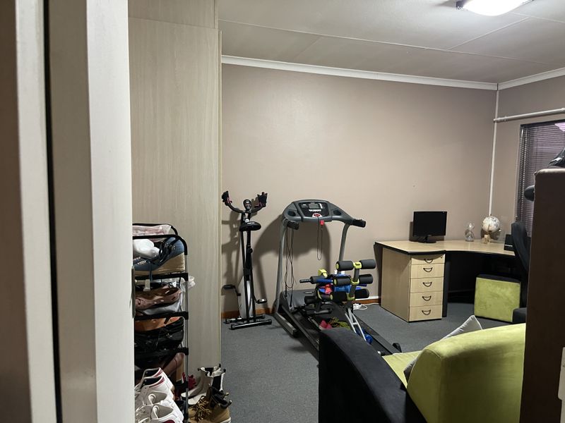 A multi-purpose bedroom featuring built-in cupboards, carpeted flooring, and window blinds, currently utilized as a home office and gym space.