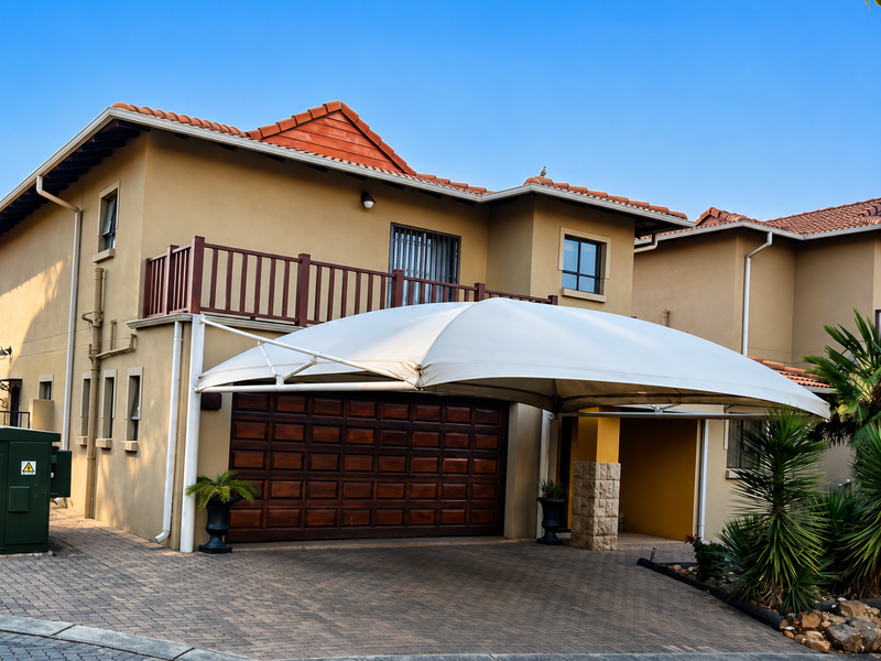 Front elevation of a double-story house featuring a paved driveway, a large shade sail carport, a wooden garage door, and an upper-level balcony.