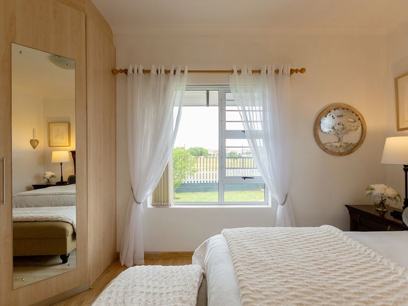 A well-maintained bedroom featuring light wood-tone built-in wardrobes with a full-length mirror, laminate flooring, and a window overlooking the garden.