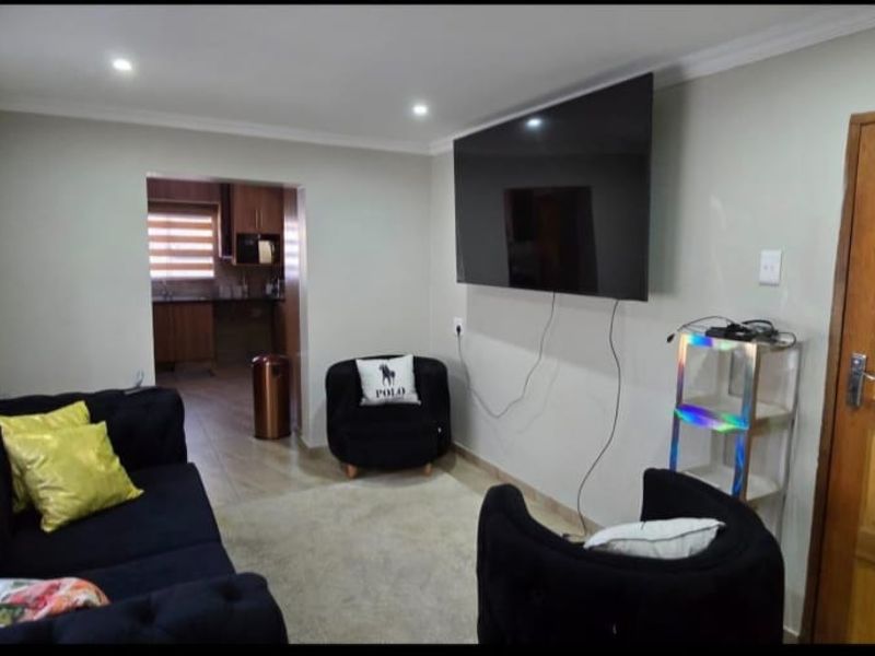 A comfortable living room featuring recessed downlights, tiled flooring, and a solid wooden door, with an open-plan flow leading into the kitchen area.