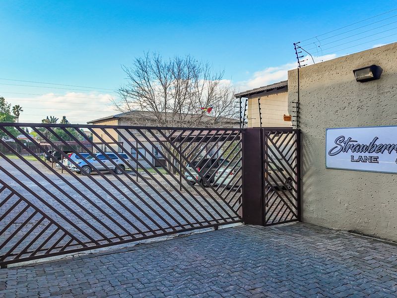 Front entrance view of the townhouse complex featuring a secure sliding gate, electric fencing, and paved driveway access.