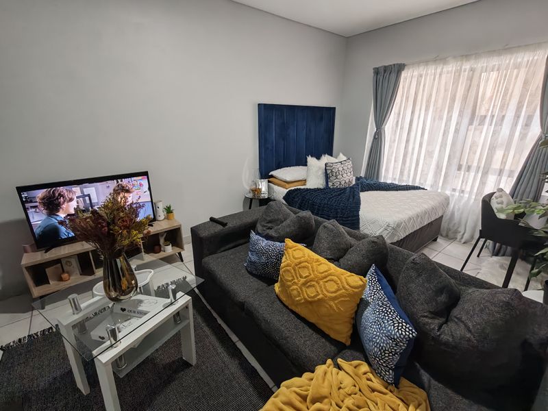 An open-plan studio apartment featuring tiled flooring and large windows with sheer curtains, providing a functional living and sleeping space.