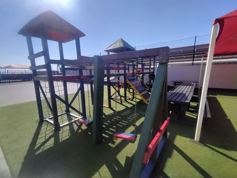 Outdoor communal playground area featuring fixed play equipment, artificial turf surfacing, and a shade structure within a secure fenced complex.