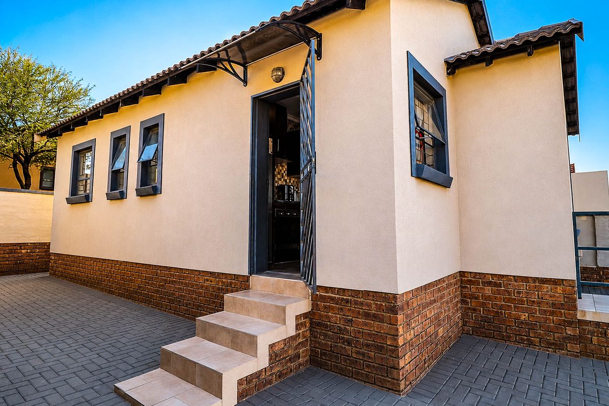 A well-maintained rear exterior view featuring a paved courtyard, brick base detailing, tiled roof, and a secure entrance with an awning.