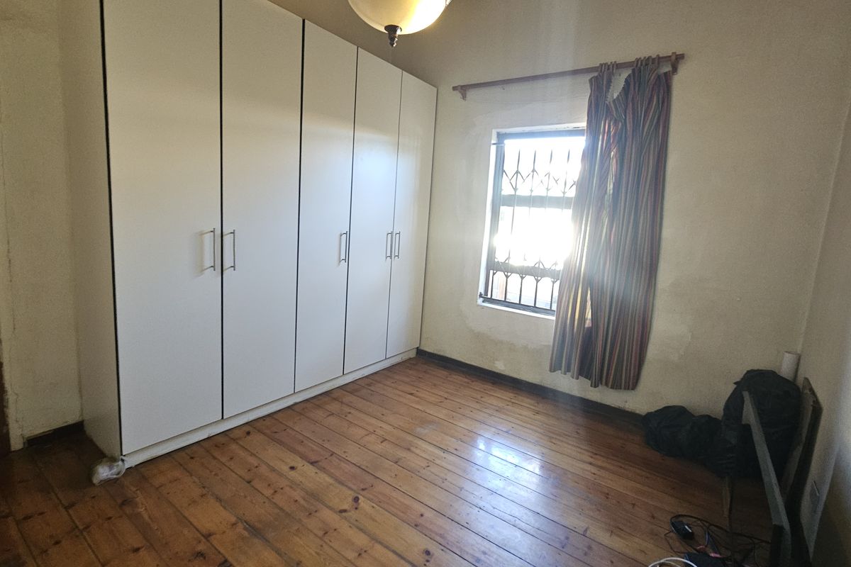 A bedroom featuring timber flooring, built-in cupboards, and a window with security bars, requiring cosmetic updates to the walls and flooring.