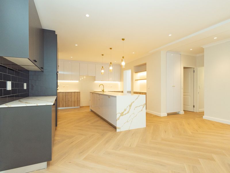 Modern, high-end kitchen featuring a large stone-topped island, herringbone timber flooring, sleek cabinetry, and elegant pendant lighting in a bright, open-plan space.
