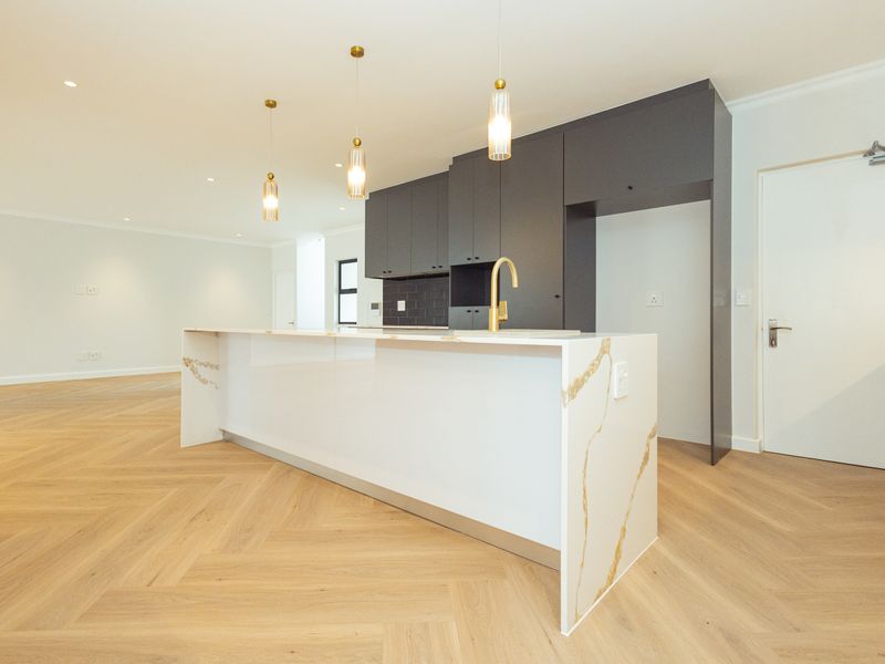 Modern open-plan kitchen featuring a stone-topped island, dark cabinetry, brass fixtures, and elegant herringbone timber flooring in a bright, spacious living area.