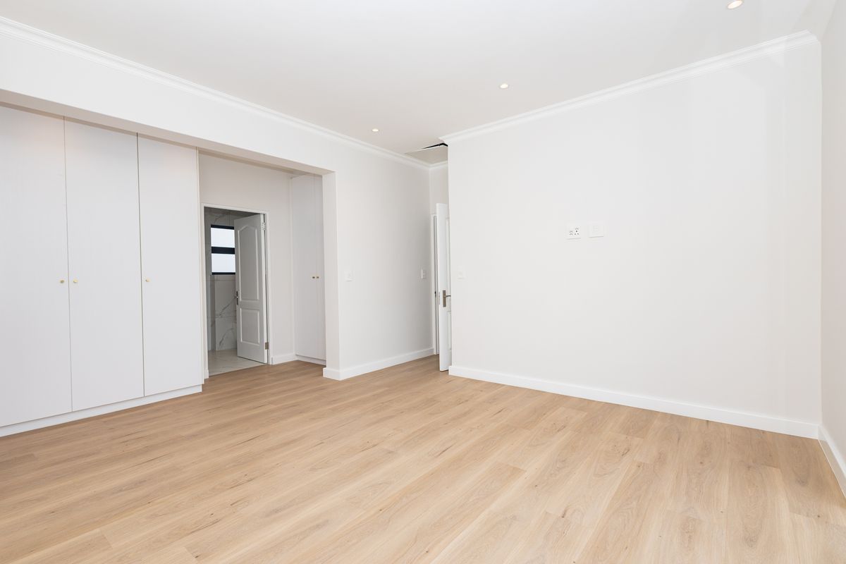 A bright, empty bedroom featuring light wood-look laminate flooring, built-in cupboards, and clean white finishes with recessed lighting.