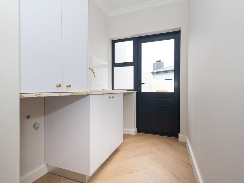 A modern, well-appointed laundry room featuring sleek white cabinetry, stone countertops, brass fixtures, and durable herringbone wood flooring with direct exterior access.