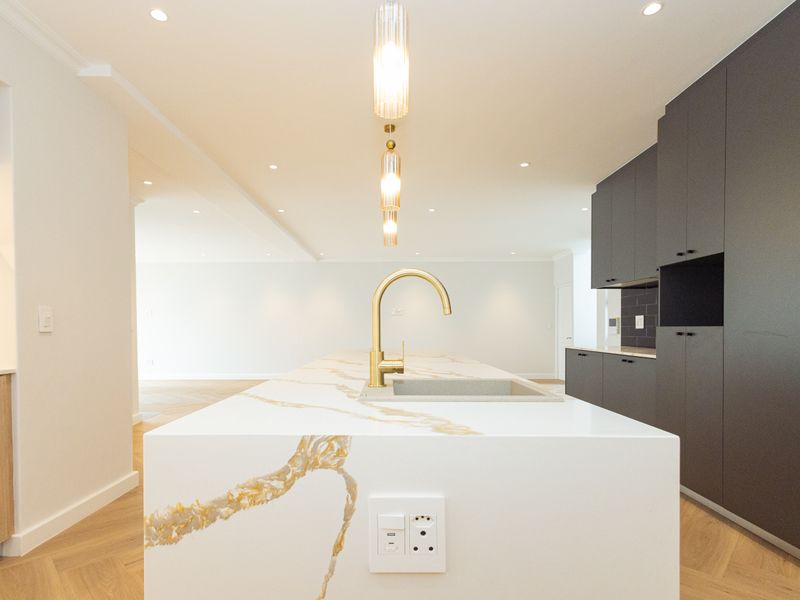 Modern kitchen featuring a large stone island with brass faucet, sleek dark cabinetry, and elegant herringbone timber flooring in an open-plan layout.