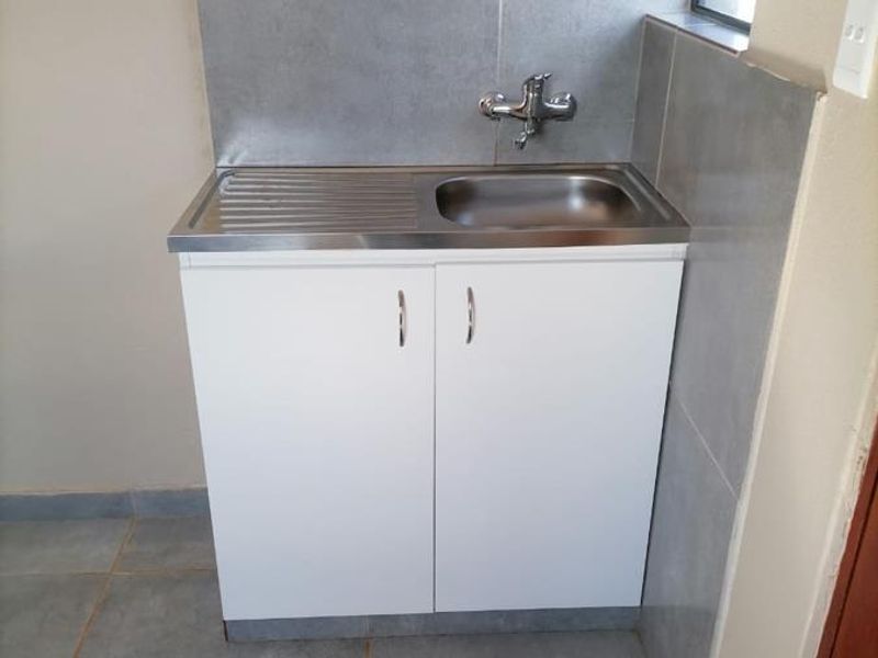 A functional laundry area featuring a stainless steel sink with mixer tap, built-in white base cabinetry, and grey tiled splashback and flooring.