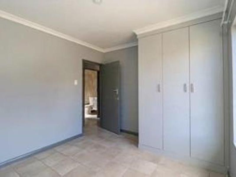 A clean, empty bedroom featuring neutral grey walls, tiled flooring, and built-in cupboards, with a doorway leading to an adjacent room.