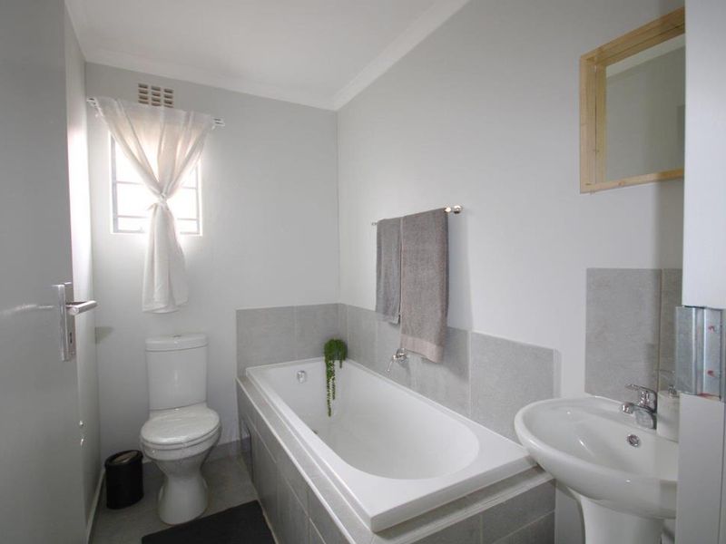 A functional bathroom featuring a built-in bathtub, pedestal basin, and toilet with neutral wall tiles and natural light from a side window.