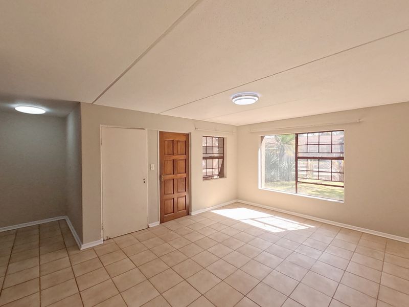 A bright, empty living area featuring neutral tiled flooring, a timber entrance door, and large steel-framed windows allowing for natural light.