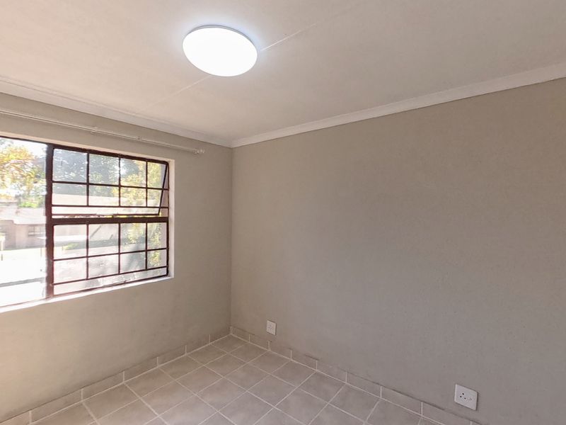 A clean, empty bedroom featuring neutral wall paint, tiled flooring, and a window fitted with security burglar bars.