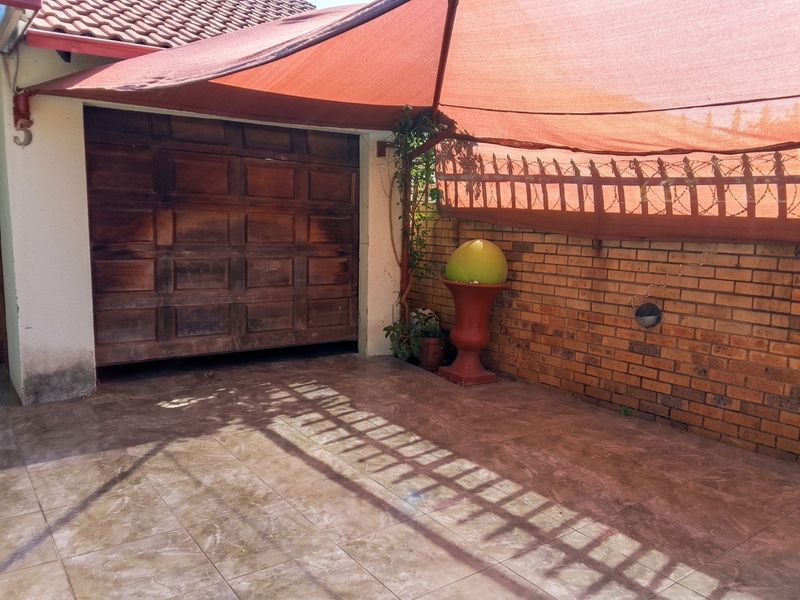 A paved garage entrance area featuring a wooden garage door, brick boundary wall with security spikes, and a protective shade sail overhead.