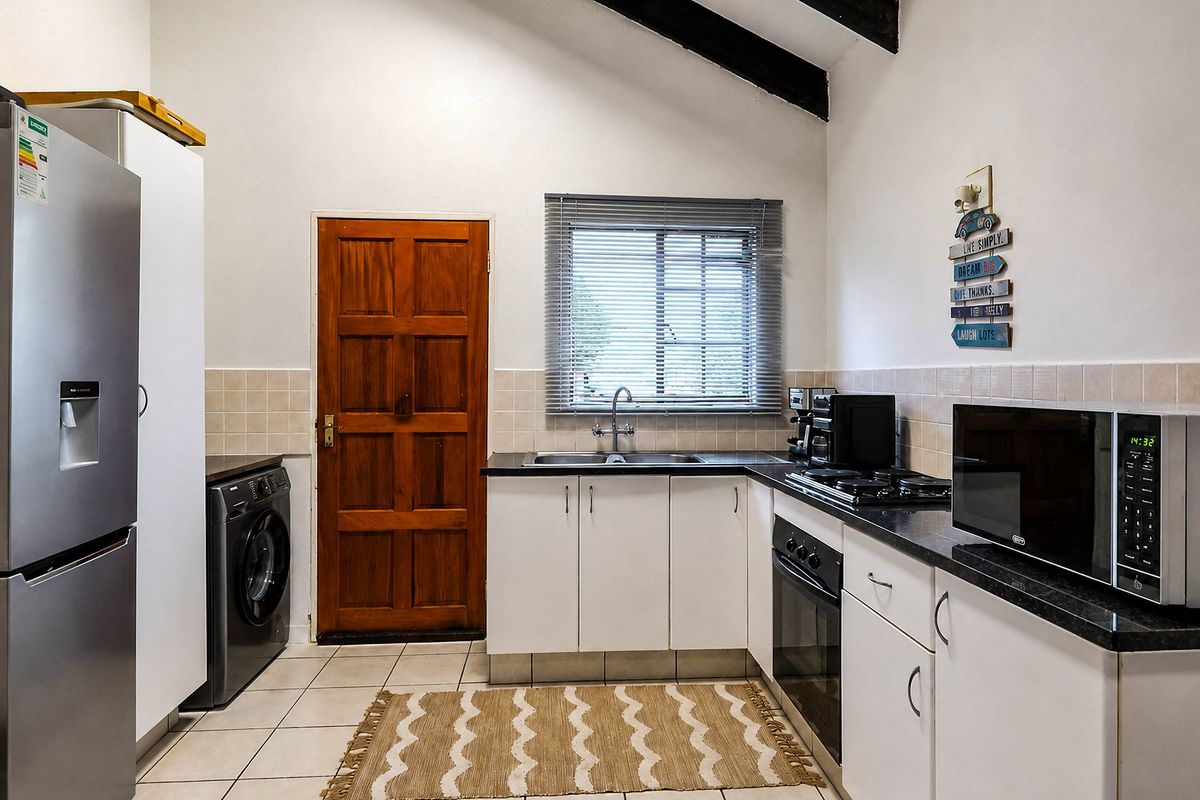 Functional kitchen featuring white cabinetry, dark countertops, built-in oven and hob, tiled flooring, and exposed timber ceiling beams with direct exterior access.