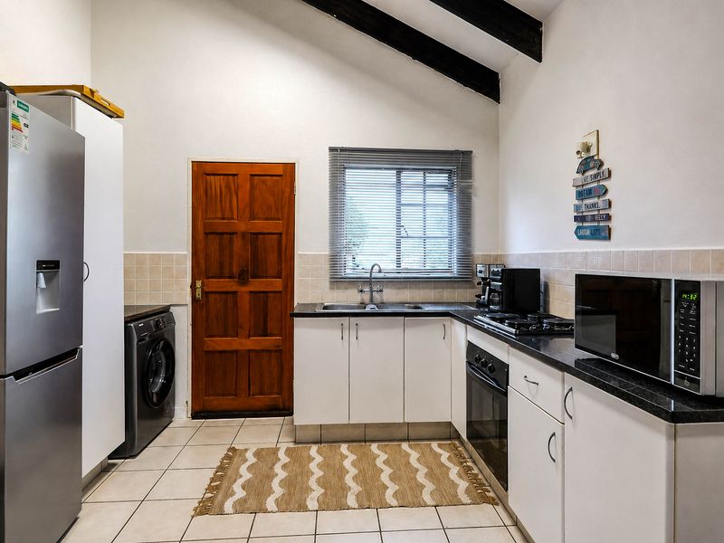 Functional kitchen featuring white cabinetry, dark countertops, built-in oven and hob, tiled flooring, and exposed timber ceiling beams with direct exterior access.