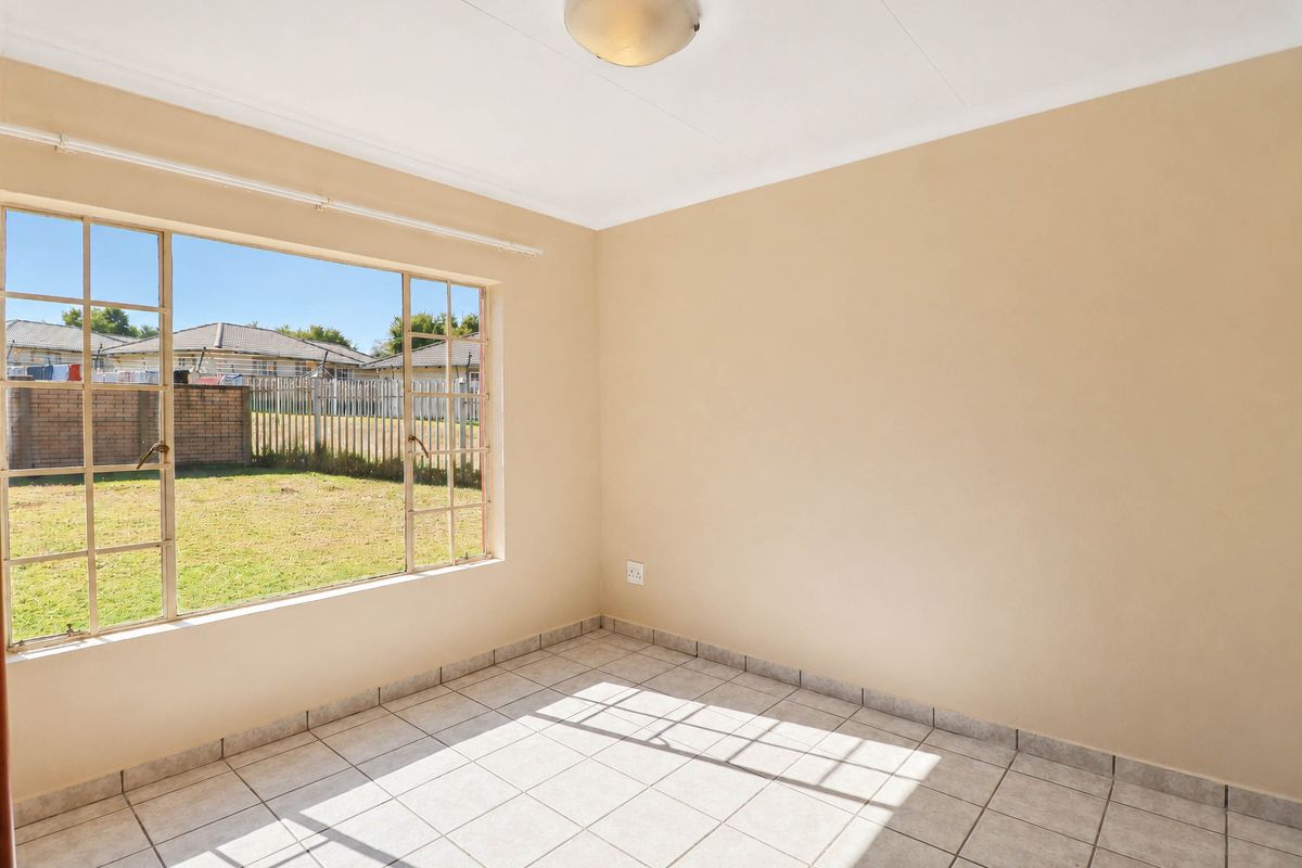 A bright, empty bedroom featuring neutral walls, tiled flooring, and large metal-framed windows overlooking the rear garden area.