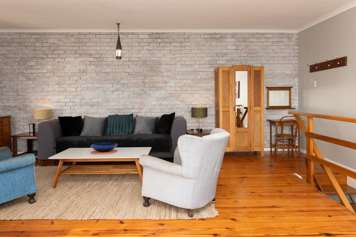 A cozy living room featuring warm timber flooring and a distinctive white-washed exposed brick feature wall, complemented by a timber staircase railing.