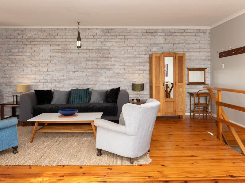 A cozy living room featuring warm timber flooring and a distinctive white-washed exposed brick feature wall, complemented by a timber staircase railing.