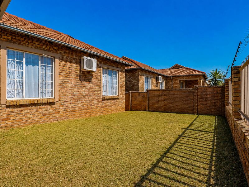 A well-maintained rear garden area featuring a face brick townhouse facade, tiled roof, and secure perimeter wall with electric fencing.