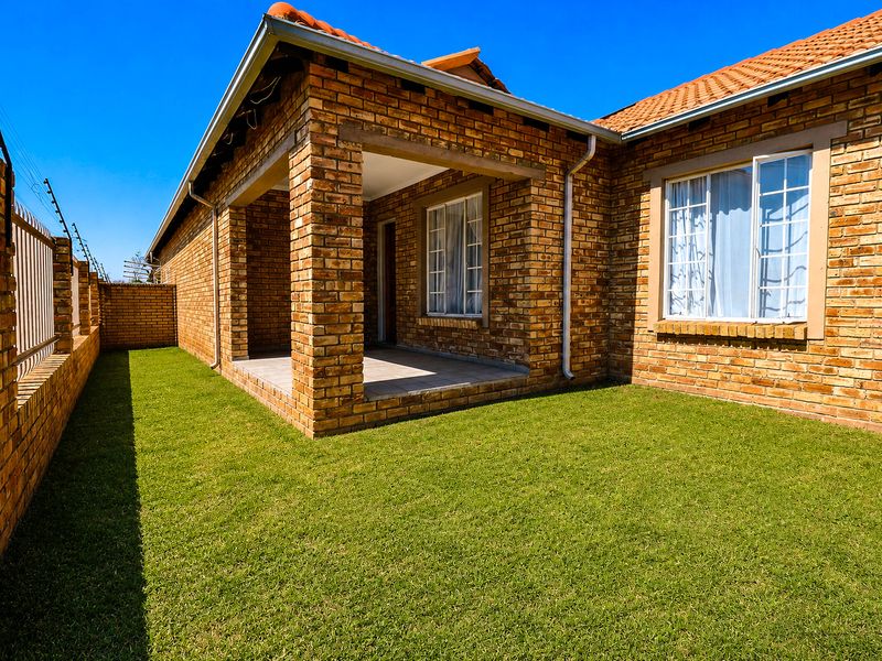 A well-maintained rear exterior view of a face brick townhouse featuring a covered patio, neat lawn, and secure perimeter wall with electric fencing.