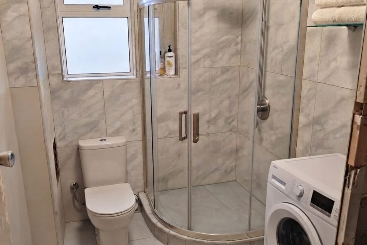 Compact bathroom featuring a corner shower enclosure, toilet, and tiled finishes, with an integrated laundry area and unfinished wall detailing near the entrance.