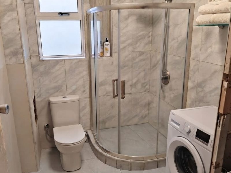 Compact bathroom featuring a corner shower enclosure, toilet, and tiled finishes, with an integrated laundry area and unfinished wall detailing near the entrance.