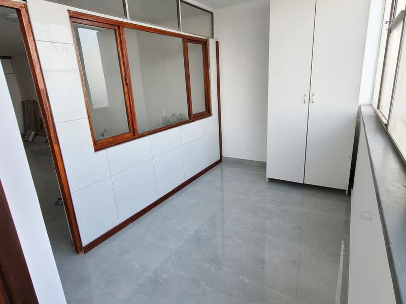 A bright, enclosed balcony or sunroom featuring modern grey tiled flooring, a built-in white cupboard, and large windows with wooden frames.
